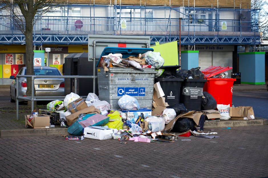 A collection of overflowing waste and rubbish bags piled beside a grey mixed paper and card recycling bin, with various cardboard boxes, plastic bags, and loose debris scattered on the paved pavement. The rubbish is situated near a metal railing that separates the sidewalk from parked cars, including a silver vehicle with a visible license plate. In the background, there are storefronts with signs and a building under renovation, covered in blue scaffolding and protective netting, indicating an urban commercial area. The scene shows an accumulation of waste that appears to have been improperly disposed of outside designated collection points, highlighting issues related to rubbish management and illegal dumping. The overall environment is an outdoor parking area adjacent to retail premises, with natural daylight illuminating the scene and light shadows cast by nearby trees and surroundings.