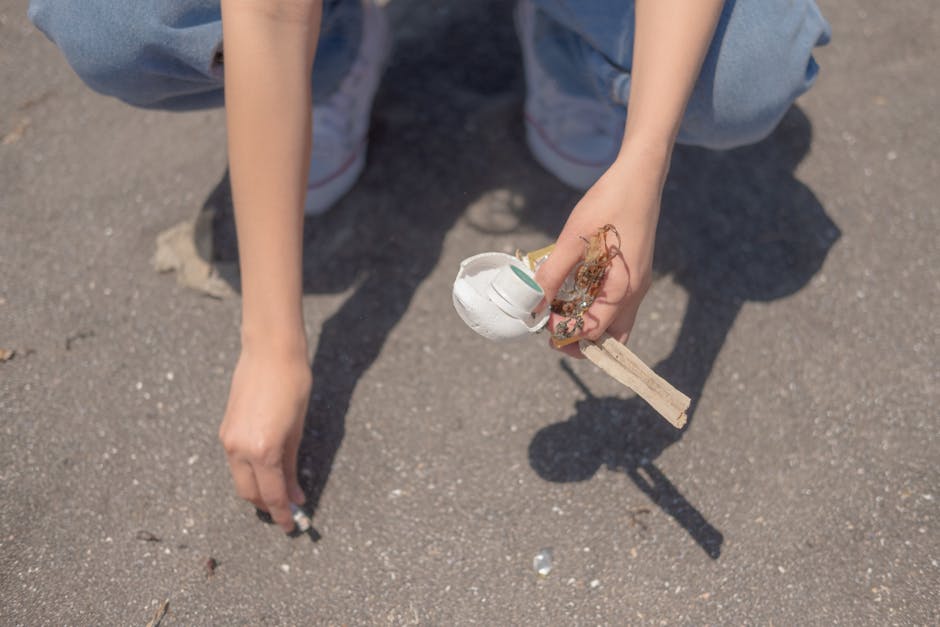 A person wearing light blue jeans and white sneakers is crouched down on an asphalt surface, holding a small, partially broken white ceramic object attached to a wooden stick or handle. The object appears to be a fragile piece of domestic or decorative ceramic, with some visible dirt or debris on it. The asphalt ground beneath them has small stones and a few scattered pieces of litter, indicating an outdoor setting. The scene suggests an act of outdoor rubbish clearing or debris collection, which could be related to private waste disposal or clean-up activities facilitated by a rubbish removal service. The natural daylight highlights the textures of the ceramic and asphalt, contributing to a clear and detailed visual description aligned with professional waste management content.