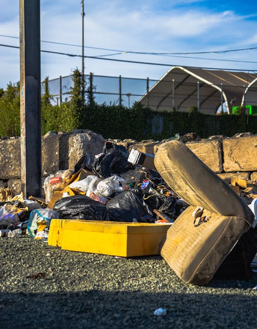 A collection of overflowing waste and rubbish bags piled beside a grey mixed paper and card recycling bin, with various cardboard boxes, plastic bags, and loose debris scattered on the paved pavement. The rubbish is situated near a metal railing that separates the sidewalk from parked cars, including a silver vehicle with a visible license plate. In the background, there are storefronts with signs and a building under renovation, covered in blue scaffolding and protective netting, indicating an urban commercial area. The scene shows an accumulation of waste that appears to have been improperly disposed of outside designated collection points, highlighting issues related to rubbish management and illegal dumping. The overall environment is an outdoor parking area adjacent to retail premises, with natural daylight illuminating the scene and light shadows cast by nearby trees and surroundings.