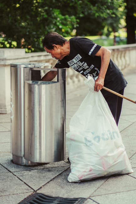 A middle-aged man with short dark hair is seen using a metal public litter bin on a paved sidewalk in a park or urban area during daylight. He is wearing a black T-shirt with white graphic text on the front and dark trousers. The man is bent over, actively depositing rubbish into the open-top, cylindrical metal bin which has a smooth, reflective surface and a rounded edge at the top. In his right hand, he holds a white plastic bag filled with refuse, while his left hand, holding a wooden stick or stick-like tool, supports the bag or assists in placing waste into the bin. The background features lush green trees and foliage, suggesting a park-like setting with natural lighting creating a sense of daytime activity. The scene is neat and orderly, highlighting common urban waste disposal practices, possibly supported by private or community-managed rubbish collection services, as evidenced by the man's engagement in proper litter disposal away from official waste collection points. The environment appears clean, with a focus on responsible rubbish handling in an outdoor public space.