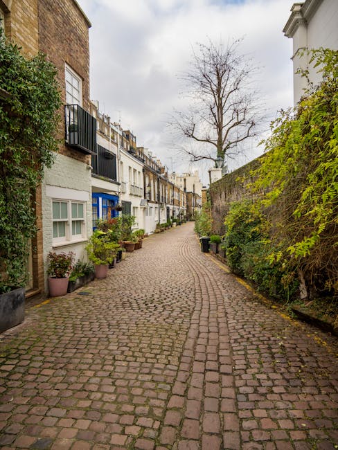 A narrow cobbled street in a residential area during daytime, lined with closely packed terraced houses featuring white and beige facades, some with small balconies and black iron railings. Potted plants and greenery are positioned along the edges of the pavement, with a tall, leafless tree standing prominently in the background against a cloudy sky. On the right side, a high stone wall covered with green foliage separates the street from private gardens or yards, while on the left, a brick building with a bay window is partially visible. The scene appears quiet and tidy, with no visible vehicles or pedestrians, and captures the typical appearance of a peaceful, historic London neighborhood that might be a candidate for private or independent rubbish removal services, emphasizing the importance of maintaining cleanliness in such charming urban environments.