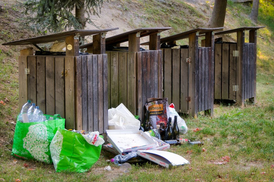 A row of four dark brown wooden outdoor rubbish and recycling bins with sloped roofs are positioned on a grassy area near sloping ground with trees in the background. The bins are made of vertical wooden planks, some with visible hinges and small metal latches on their doors. In front of the bins, various discarded items are scattered across the grass, including large green shopping bags filled with plastic bottles and other waste, a white pizza box with food remnants, empty glass bottles, and crushed plastic bottles. The scene suggests an informal disposal area outside, with the surrounding environment consisting of natural greenery, trees, and uneven terrain. This setting is indicative of private or independent waste disposal, possibly in a park or woodland edge, aligning with services like rubbish removal for on-site clearance or alternative waste handling.