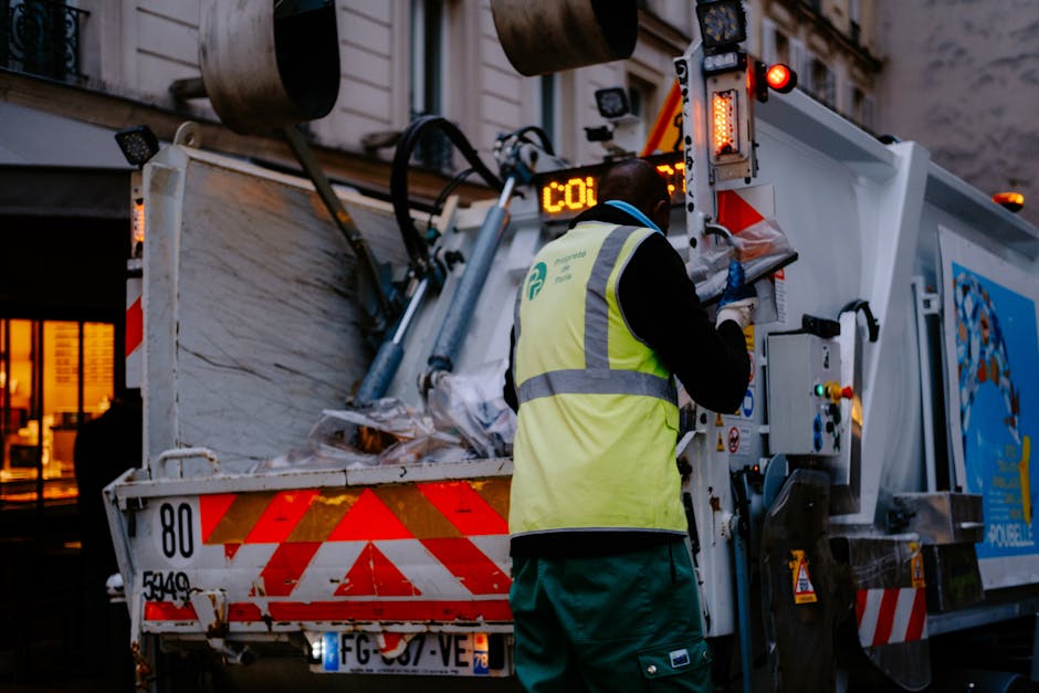 A worker wearing a high-visibility yellow vest with reflective silver stripes is operating a waste collection vehicle on a city street during the evening. The worker's back is turned, and they are using a control panel on the side of the truck, which has a white body with red reflective stripes and a blue logo on the side. The rear of the vehicle is open, revealing a large, overflowing hopper filled with mixed waste materials, including crumpled paper, plastic, and other refuse. The surface of the waste appears crumpled and compressed, with a mixture of textures and colors such as white, black, and transparent. In the background, there are illuminated windows of a nearby building, suggesting an urban environment. The scene is lit with ambient street lighting, creating a contrast between the bright artificial lights and the darker surroundings, highlighting the operational activity of waste collection often carried out during off-peak hours, supporting alternative waste handling methods outside typical municipal rubbish collection.