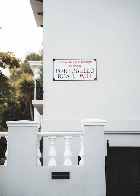 A white residential building exterior featuring a decorative white balustrade with three evenly spaced balusters and a small rectangular mail slot beneath it. On the building's smooth white wall, there is a rectangular street sign with a black border, white background, and red text indicating it is located on Portobello Road in the Royal Borough of Kensington and Chelsea, with the postal code W11. The sign is mounted close to the top left corner of the wall, and part of a balcony with black wrought iron railing is visible extending from the upper floor on the left side. In the background, there are leafy trees with green and yellow foliage, suggesting a suburban area. The scene is illuminated by natural daylight, with a soft, neutral atmosphere. The overall impression is of a tidy, well-maintained urban street scene, potentially involving private property, consistent with independent waste collection or on-site rubbish management practices in the area.