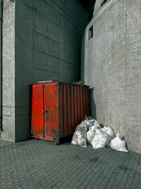 A metal rubbish skip with a weathered, rusty red finish is positioned against a building corner on a paved surface. The skip is rectangular with vertical ridges along the sides and a hinged door on one end. Surrounding the base of the skip are several tightly tied white and clear plastic bags filled with waste, some bulging from the weight, indicating general household or commercial rubbish. The background includes a dark, smooth wall on the left with horizontal paneling and a grey, textured wall on the right, possibly concrete. A small window or vent is visible high on the right wall, and the scene appears to be outdoors, possibly on a service access area or alleyway. The lighting is diffuse, suggesting an overcast sky, with no direct sunlight or shadows present. This scene exemplifies a typical private waste collection point, often used for on-site rubbish clearing or alternate disposal methods aside from council collection services, aligning with the context of independent rubbish removal services near South Kensington station.
