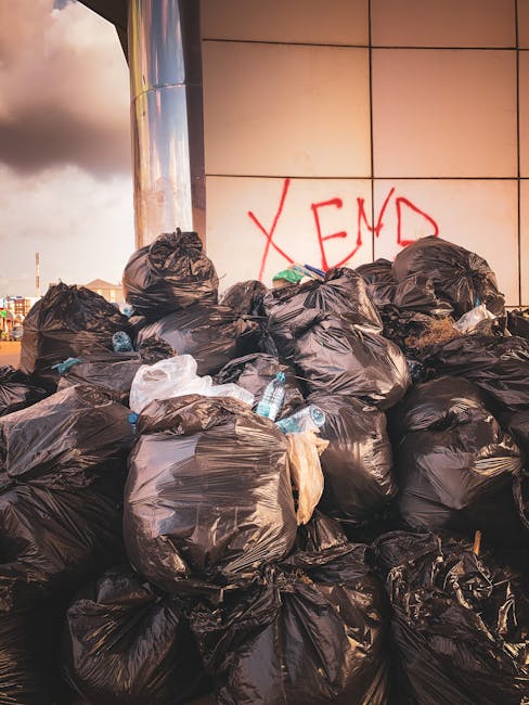 A large collection of black plastic garbage bags piled against a tiled wall in an outdoor urban environment, with some water bottles and miscellaneous waste protruding from the bags. The wall behind features graffiti with the message 'XEND' spray-painted in red. The scene is illuminated with natural light, casting soft shadows on the bags and highlighting their shiny, reflective surfaces. The background shows part of a cityscape under an overcast sky, indicating an area of informal waste disposal or unsanctioned rubbish accumulation, consistent with private rubbish handling or on-site clearance activities related to alternative waste management options or independent collection services.
