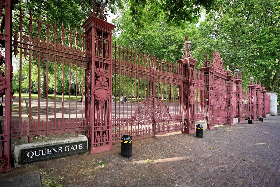 A long section of ornate wrought iron fencing painted in a deep reddish-pink color, featuring decorative vertical bars with pointed finials at the top and intricate embossed detailing on the supporting pillars. The fence extends across the image, with several tall, matching pillars topped with decorative lantern-style lights and finials positioned at regular intervals. To the left at the base of the fence, a rectangular black sign with white lettering reads 'QUEENS GATE.' The ground beneath the fence is paved with cobblestones, and small black bollards with reflective yellow stripes are aligned along the front edge. Behind the fence, lush green trees with dense foliage partially obscure the view into the adjacent park or estate grounds, creating a natural canopy overhead. The lighting appears natural, suggesting daytime, with diffused sunlight filtering through the leaves. The scene conveys a historic or classical aesthetic, relevant to the surrounding area or property, and highlights the importance of maintaining or potentially removing old fencing structures as part of exterior property management.