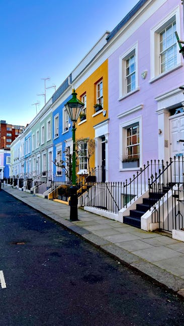 A row of colourful Victorian terraced houses along a paved street, with each property painted in pastel shades of blue, yellow, and lavender. The houses feature white framed sash windows, some with flower boxes, and small front gardens enclosed by low black iron railings. A traditional black lamp post with a green top is positioned on the pavement in the foreground. The steps leading up to the front doors are black with white risers, and a narrow street with slight incline runs parallel to the row of houses. The scene is illuminated by natural daylight, with a clear sky above, and television antennas are visible atop some of the buildings. This setting exemplifies private residential properties that may be serviced by independent rubbish removal companies, balancing environmental aesthetics with waste management needs in a typical urban neighbourhood.