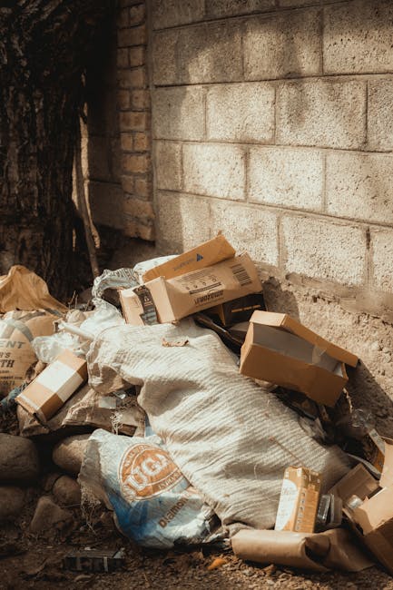 A pile of discarded cardboard boxes and flattened parcels, partially wrapped in white and brown packaging materials, is situated at the base of a weathered brick wall in an outdoor setting. The cardboard boxes vary in size and some have printed labels and barcodes visible on the surface, indicating they are used packaging waste. There are also crumpled brown paper and clear plastic wrapping around the debris, suggesting they are remnants from recent deliveries or packaging. The pile is positioned directly against a rough-textured brick wall constructed of light grey and beige bricks, with visible mortar joints. To the left, a section of dark tree bark is partially visible, indicating proximity to a tree or natural environment. The ground beneath the rubbish comprises loose stones, dirt, and small debris, with soft, natural lighting highlighting the textures and materials of the waste and wall. This scene exemplifies typical clutter associated with private waste disposal or collection of packaging materials, aligning with services offered by rubbish removal companies like Rubbish Removals South Kensington.