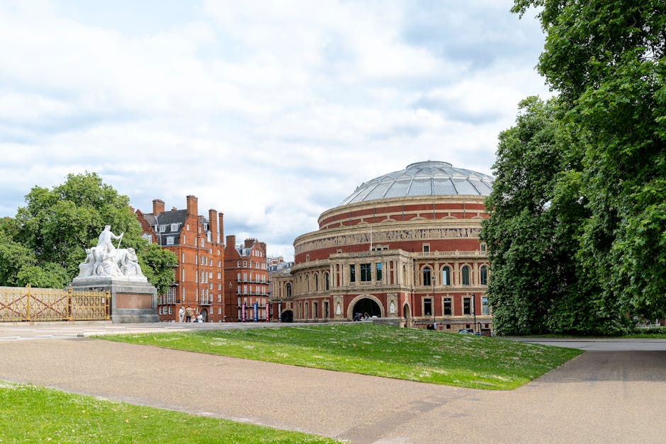 A wide shot of the Royal Albert Hall, a large circular building with a prominent domed roof made of metal and glass, situated in a park with well-maintained grassy areas and paved walkways. Surrounding the hall are tall, leafy green trees and a white marble sculpture depicting figures seated and standing, positioned on a stone plinth to the left side of the image. In the background, residential brick buildings with chimneys can be seen behind the trees. The sky is partly cloudy with patches of blue, providing natural daylight that illuminates the scene evenly. The park appears peaceful, with no visible litter or debris, illustrating a setting where outdoor waste management and occasional rubbish removal might be required, such as clearing fallen leaves or refuse after events or public gatherings. The overall scene emphasizes a clean, organized outdoor environment near a historic cultural landmark, aligning with the context of private waste handling or waste management services like those offered by rubbishremovalsouthkensington.org.uk in the area.