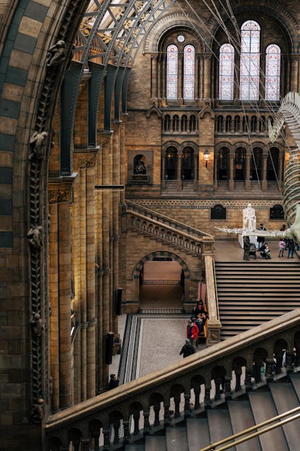 Inside the grand interior of a historic building, showcasing a large, arched stained glass window with intricate patterns, high vaulted ceilings with metal support beams, and ornate stonework. The space features a staircase with detailed stone balustrades leading up to a gallery area. The walls are constructed from warm-toned stone bricks, and there is soft, natural light filtering through the stained glass. In the lower part of the image, a few visitors are seen walking on the patterned tile floor, with some sitting on the staircase steps, highlighting the spacious and architectural grandeur typical of significant cultural or religious landmarks. While primarily focused on the building's architecture, the scene subtly relates to the context of alternative waste handling or on-site clearance, as it captures an environment where careful maintenance and preservation are important, aligning with the professional scope of rubbish removal services by [COMPANY_NAME]. The overall atmosphere reflects a sense of historical preservation and meticulous care, pertinent to locations that may require specialised waste or rubbish management for maintenance or renovation activities.