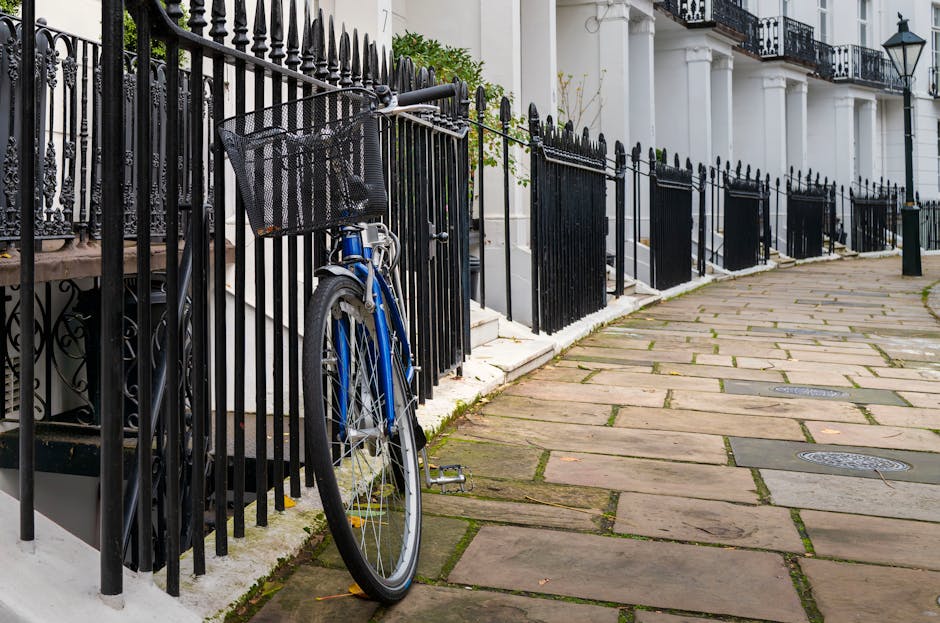 A blue bicycle with a black front basket is secured and leaning against a black wrought iron fence bordering a residential building. The fence is composed of vertical bars with pointed finials on top, situated along a sidewalk paved with irregularly shaped sandstone slabs. The background features white-painted terraced houses with decorative cornices, black wrought iron railings on the balconies, and a traditional street lamp attached to one of the buildings. The scene is illuminated by natural daylight, with shadows cast on the pavement. The fence runs parallel to the building's entrance steps, which are partially visible, and the environment appears clean and well-maintained, reflecting an urban street scene where private property boundaries are marked by ornamental fencing. The overall setting aligns with residential areas in South Kensington where independent waste management may take place, although no waste or rubbish is visible in the image.