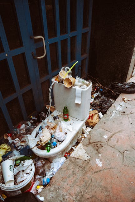A white ceramic toilet that is no longer in use is situated outdoors against a dark, metal gate with vertical bars and a curved handle. The toilet is surrounded by a significant accumulation of miscellaneous rubbish, including crumpled paper, plastic bottles, and food packaging, scattered on the ground around it. A small green glass bottle and a few disposable cups are placed on top of the toilet tank, with one cup containing a plastic straw. To the left of the toilet, a plastic bucket or container holds more waste, including paper and plastic items. The ground surface is a combination of cracked concrete and dirt, with some wet patches and scattered debris. The environment appears to be an outdoor alley or back area, with the debris indicating an informal or neglected waste disposal spot, possibly related to private rubbish accumulation rather than a regulated collection point. The scene illustrates issues with improper waste management and the need for regular rubbish removal service, such as that provided by rubbishremovalsouthkensington.org.uk, to prevent clutter and maintain cleanliness in such outdoor spaces.