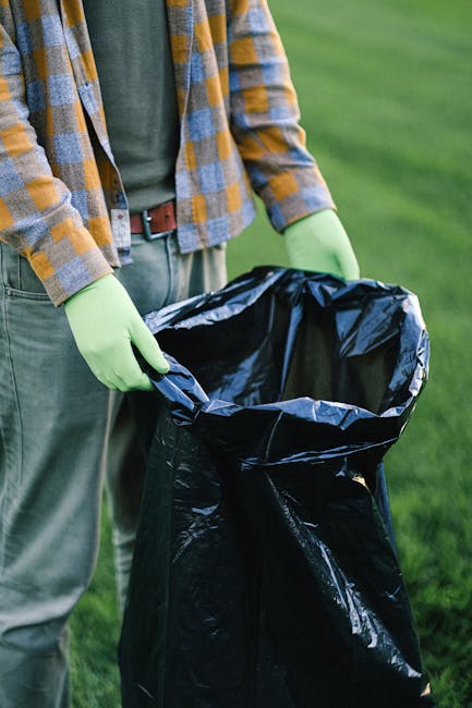 A pile of mixed waste and cardboard boxes is accumulated against a weathered brick wall outside, close to a large tree trunk visible on the left side of the image. The waste includes flattened and crumpled cardboard packaging, some with printed labels and barcodes, alongside plastic bags and a white fabric sack. The debris rests on uneven ground with small stones and dirt, indicating an outdoor setting such as a driveway or alleyway. The cardboard boxes appear to be partially open, with some torn or damaged, and the surfaces show signs of dirt and outdoor exposure. The brick wall in the background has a rough, textured surface with visible mortar joints, indicating a sturdy structure, while the environment suggests an informal collection point for waste awaiting disposal or removal through private rubbish collection services. The scene emphasizes the importance of proper waste management and highlights the potential obstruction to access for waste removal in this specific area.